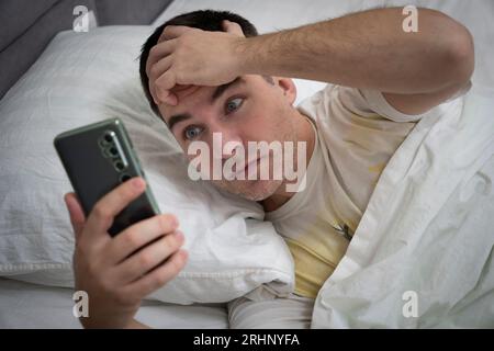 Homme surpris d'entendre la sonnerie de l'alarme de smartphone, se trouve dans le lit blanc. Réveillez-vous un gars choqué tard pour le travail le matin. Surprise du message sur smar Banque D'Images