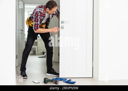 Installation d'une serrure sur la porte d'entrée avant en bois. Portrait d'un jeune ouvrier de serrurier en uniforme bleu en installant le bouton de porte. Professionnel Banque D'Images