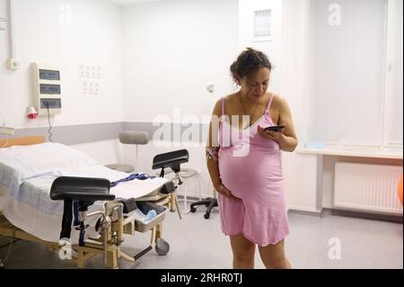 Femme enceinte, mère accoucheuse en travail, vêtue d'une chemise de nuit rose, dans une chambre d'hôpital, tient son ventre et compte ses contractions, à l'aide d'un mobi Banque D'Images