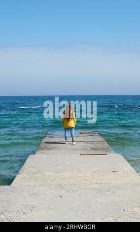 Jeune femme aux cheveux longs et portant une veste coupe-vent jaune marchant sur la jetée entourée par la mer Banque D'Images