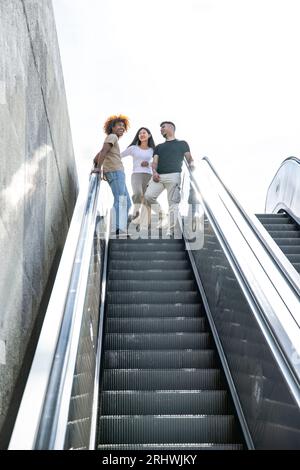 Groupe multiethnique d'amis riant et descendant un escalator extérieur. Trois jeunes gens joyeux souriants et l'air détendus marchant vers le bas de la Banque D'Images