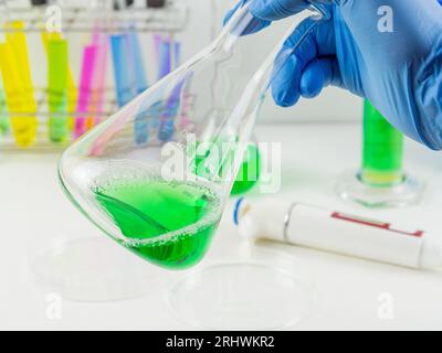 Gros plan d'une main portant des gants tenant un erlenmeyer avec un échantillon liquide vert dans un laboratoire. Banque D'Images