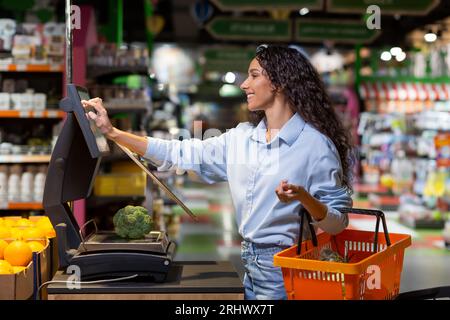 La jeune femme acheteuse dans le supermarché utilise des balances en libre-service, souriante femme hispanique pèse les fruits et légumes dans un grand magasin. Banque D'Images