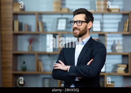 Portrait de patron mature réussi, homme d'affaires senior dans des lunettes et costume d'affaires regardant loin et souriant, homme aux bras croisés travaillant à l'intérieur d'un immeuble de bureaux moderne. Banque D'Images