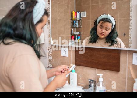 Fille asiatique dans la bande de cheveux propre des dents dentiers dans l'évier en face du miroir dans la salle de bain, concept de routine matinale Banque D'Images