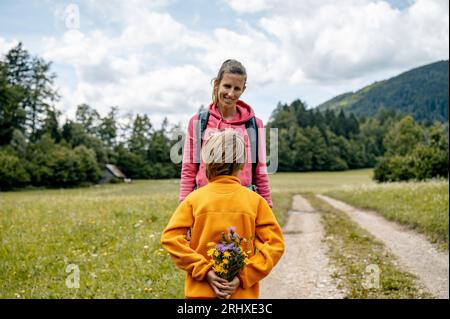 Vue de derrière d'un garçon en bas âge cachant bouquet de fleurs cueillies à la main derrière son dos pour le donner à sa mère. Dehors dans la belle nature d'été f Banque D'Images