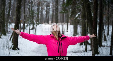 Heureuse jeune femme sereine en veste d'hiver rose debout avec ses bras écartés au milieu de la forêt d'hiver couverte de neige. Banque D'Images