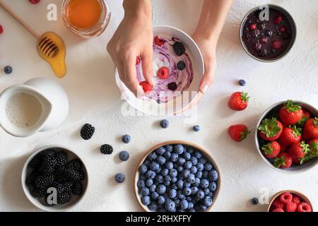 Vue de dessus des mains de femme de récolte préparant le petit déjeuner avec bol avec yaourt naturel et framboises, bleuets, fraises et mûres près des bols Banque D'Images