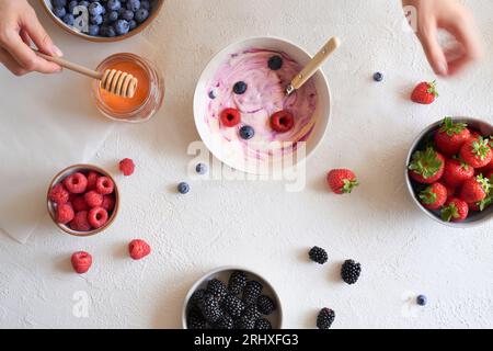 Vue de dessus des mains de femme de récolte préparant le petit déjeuner avec bol avec yaourt naturel près des bols avec du miel et différents fruits frais placés sur la table Banque D'Images