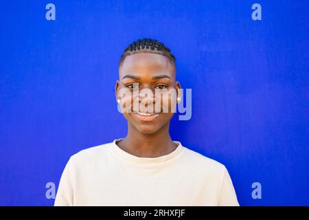 Portrait d'un adolescent afro-américain confiant en tresses et chemise blanche debout sur fond bleu tout en souriant et en regardant la caméra Banque D'Images