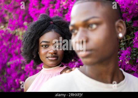 Couple d'adolescents afro-américains avec coiffure afro dans des vêtements décontractés tout en se tenant ensemble regardant la caméra dans le parc avec fleurs violettes en fleurs Banque D'Images