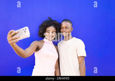 Portrait d'un couple d'adolescents afro-américains positif avec coiffure afro prenant selfie ensemble sur smartphone tout en se tenant sur fond bleu Banque D'Images