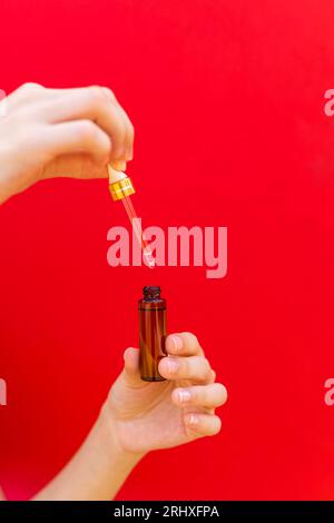 Cultivez la femelle anonyme tenant le verre ambré avec le sérum cosmétique et la pipette sur fond rouge Banque D'Images