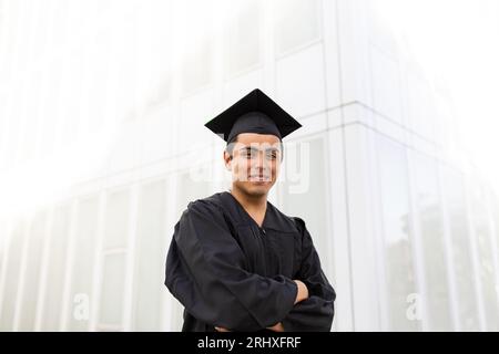 Portrait de caucasien confiant beau jeune homme portant une robe de graduation noire et une casquette debout avec les bras croisés contre le mur blanc Banque D'Images