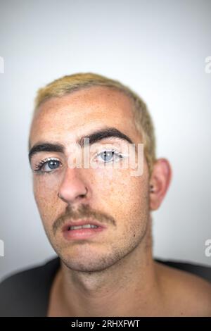 Portrait de jeune homme non rasé avec les cheveux courts moustache yeux bleus regardant la caméra dans une pièce floue contre le mur blanc à la lumière du jour Banque D'Images