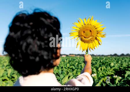 Vue arrière de la personne anonyme tenant beau tournesol jaune mûr frais avec le visage heureux contre le ciel clair pendant l'été à la ferme Banque D'Images