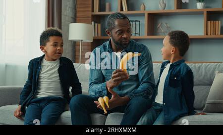 Famille afro-américaine à la maison père célibataire homme avec deux enfants garçons papa parlant avec des enfants donner bananes fruits vitamines parler expliquer enseigner à Banque D'Images