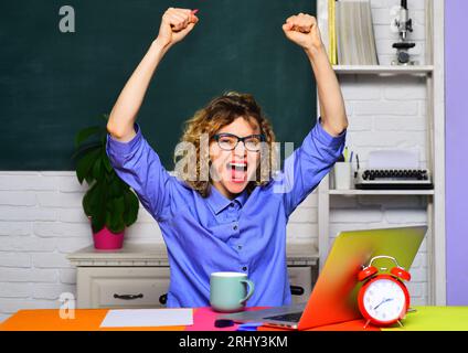 Fille étudiante excitée avec les mains levées en classe. Réussite. Heureux étudiant sur le campus universitaire. Enseignante souriante assise à table avec ordinateur portable Banque D'Images