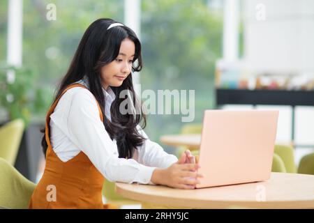 Belle adolescente asiatique avec ordinateur portable. Jeune femme avec les cheveux longs travaillant sur l'ordinateur dans le restaurant. Étudiant faisant ses devoirs. Banque D'Images