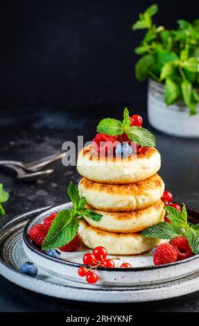 Crêpes épaisses au fromage cottage aux myrtilles, framboises et groseilles rouges, décorées de menthe fraîche sur une assiette grise, délicieux petit déjeuner. Tabl. Noir Banque D'Images