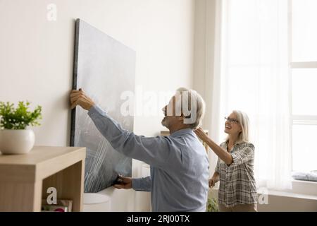 Joyeux couple senior décorant l'appartement, accrochant une grande photo Banque D'Images