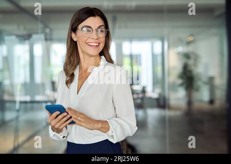 Heureuse femme d'affaires mûre souriante tenant le téléphone regardant loin dans le bureau. Banque D'Images