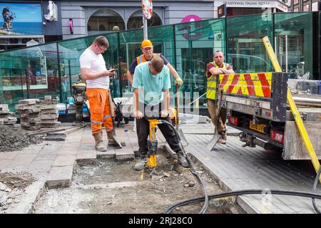 4 ouvriers réparant la chaussée, dont un travaillant sur une perceuse routière, un appuyé sur le camion, un sur son téléphone et un appuyé sur sa pelle, Glasgow, Banque D'Images