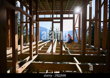 Menuisier construisant cadre en bois, maison de deux étages près de la forêt dans la soirée. Homme barbu martelant des clous avec un marteau au coucher du soleil. Concept de construction moderne respectueuse de l'environnement. Banque D'Images