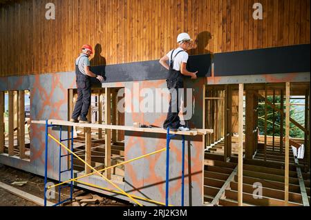 Charpentiers construisant une maison encadrée en bois. Deux hommes ouvriers claviant la façade de la maison avec des panneaux de particules de ciment, les fixant avec des tournevis. Concept de construction écologique moderne. Banque D'Images