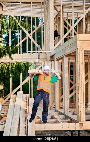 Bâtiment de charpentier maison à ossature en bois près de la forêt. Homme tenant une grande poutre sur son épaule, vêtu de vêtements de travail et casque. Idée de construction moderne et durable. Banque D'Images