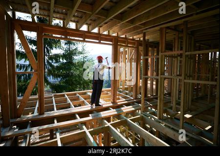 Menuisier construisant un cadre en bois, maison de deux étages près de la forêt. Homme barbu martelant des clous avec marteau, vêtu d'un casque de protection et d'une combinaison de travail. Concept de construction écologique moderne. Banque D'Images