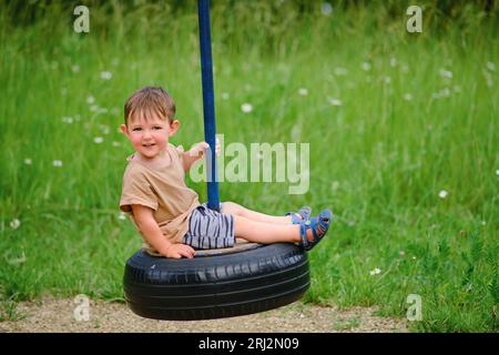 Un enfant souriant aime jouer sur un pneu de voiture transformé en balançoire. Un bébé heureux tourne sur une balançoire faite d'un pneu d'une roue de voiture. Enfant âgé de deux ans YE Banque D'Images