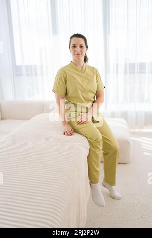 Portrait vertical pleine longueur de jolie femme masseuse médecin en uniforme assis sur un canapé blanc regardant la caméra avec expression amicale Banque D'Images