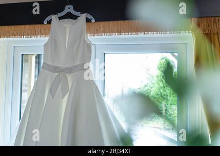Une robe de mariée blanche est suspendue à un porte-vêtements devant une fenêtre lumineuse, éclairée par une lumière chaude et naturelle Banque D'Images