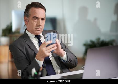 Homme d'affaires mûr sérieux vérifiant les messages texte et les notifications sur smartphone Banque D'Images