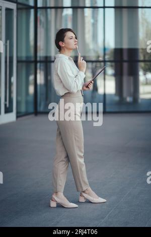 Jeune femme d'affaires caucasienne mignonne avec une tablette et un stylet dans ses mains a pensé à des projets d'affaires prometteurs. Tir long, vertical Banque D'Images
