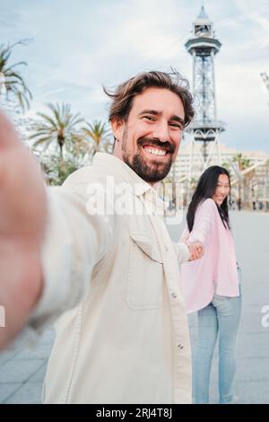 Vertical. Beau jeune homme adulte marchant et prenant un portrait selfie tenant la main de sa petite amie dans la ville sur une date. Vrai couple joyeux ayant du plaisir ensemble avec vitalité sur le voyage de week-end. Photo de haute qualité Banque D'Images