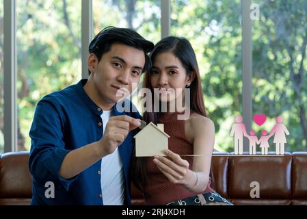 Jeune couple asiatique assis sur un canapé confortable, mettant des pièces d'argent dans une boîte à pièces en bois en forme de maison, laissant entendre les rêves d'un avenir idyllique. Banque D'Images