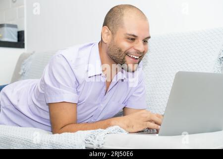 Homme riant avec chauve et barbe travaillant à l'ordinateur à l'intérieur à la maison au canapé Banque D'Images