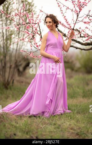 Femme fleur de pêche. Heureuse femme en robe rose marchant dans le jardin des arbres de pêche en fleur au printemps Banque D'Images