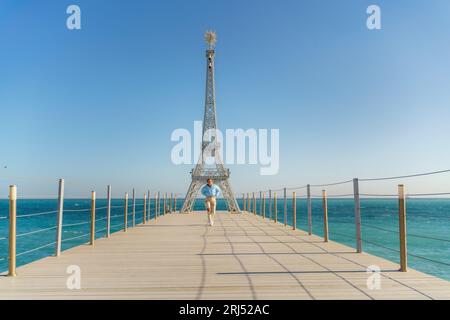 Grand modèle de la Tour Eiffel sur la plage. Une femme marche le long de la jetée vers la tour, portant une veste bleue et un Jean blanc. Banque D'Images