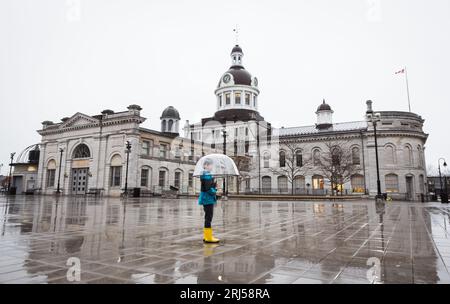 Petit garçon portant des vêtements de pluie tenant un parapluie sur une place de ville pluvieuse. Banque D'Images
