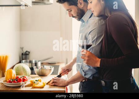 Le jeune homme prépare le déjeuner pour sa petite amie dans la cuisine pendant qu'elle boit un verre de vin Banque D'Images