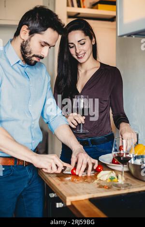 Le jeune homme prépare le déjeuner pour sa petite amie dans la cuisine pendant qu'elle boit un verre de vin Banque D'Images