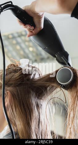 Gros plan des mains de coiffeurs séchant de longs cheveux blonds avec sèche-cheveux et brosse ronde Banque D'Images