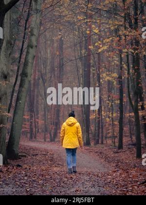 Une femme portant un imperméable jaune marchant dans une zone boisée, entourée d'un feuillage luxuriant en automne Banque D'Images