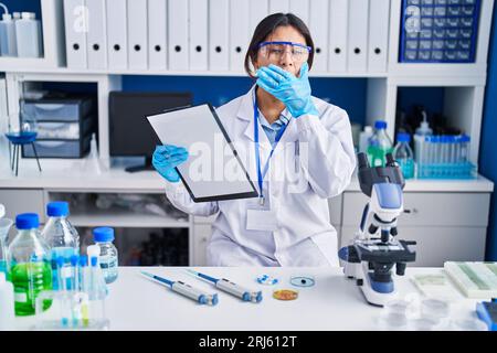 Jeune femme hispanique travaillant au laboratoire scientifique ennuyé bâillant fatigué couvrant la bouche avec la main. agité et somnolence. Banque D'Images