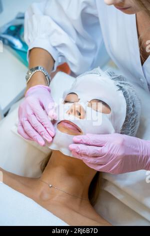 Cosmétologue portant des gants en caoutchouc appliquant le masque de feuille sur le visage de la femme. Banque D'Images