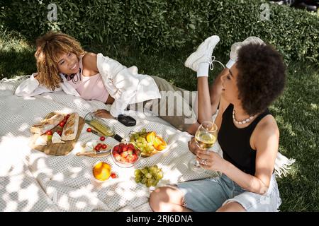 femme afro-américaine avec verre de vin parlant à la petite amie heureuse près des collations sur le pique-nique d'été Banque D'Images