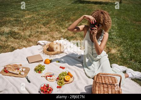 femme afro-américaine prenant la photo sur l'appareil photo vintage près des fruits et légumes, pique-nique d'été Banque D'Images